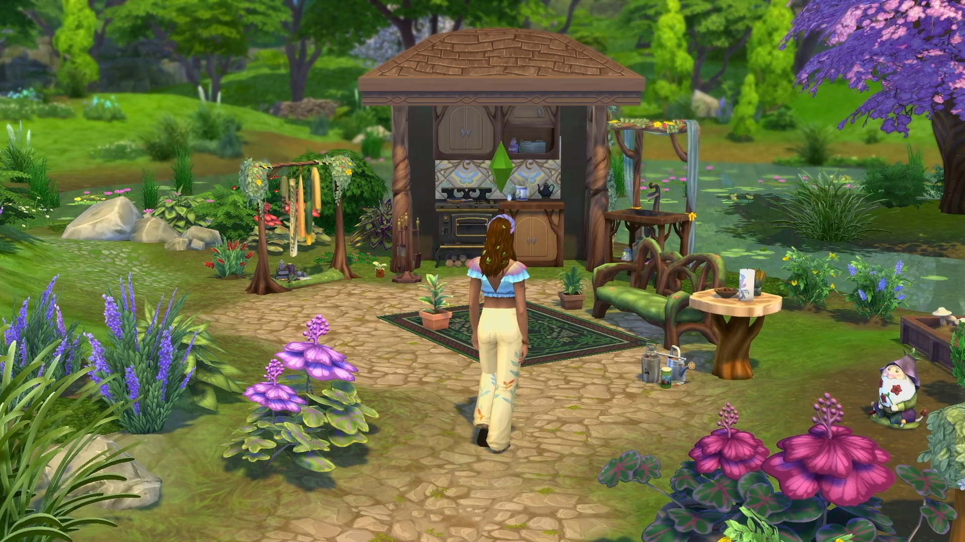 A Sim walks toward a rustic, whimsical outdoor kitchen surrounded by colorful flora and forest scenery.